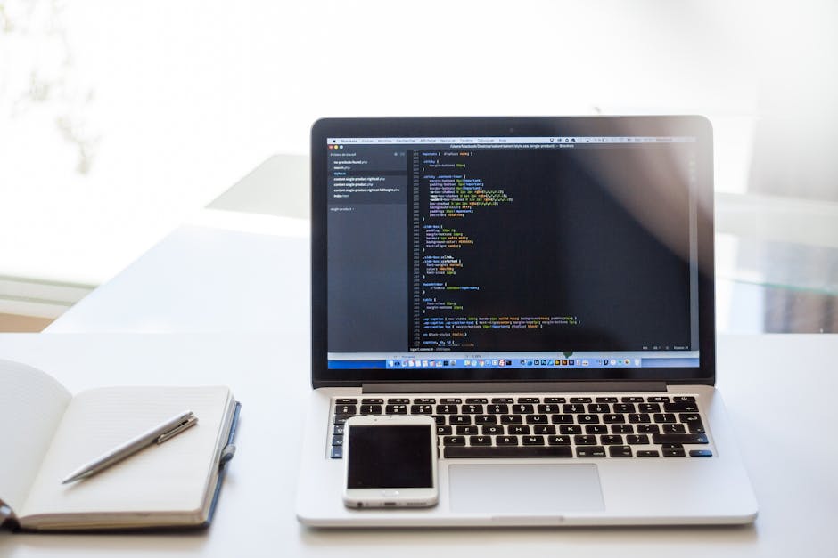 A close-up view of a clean, well-organized workspace featuring a silver laptop displaying lines of colorful code on a dark background, placed on a white desk. To the left of the laptop, there's an ope